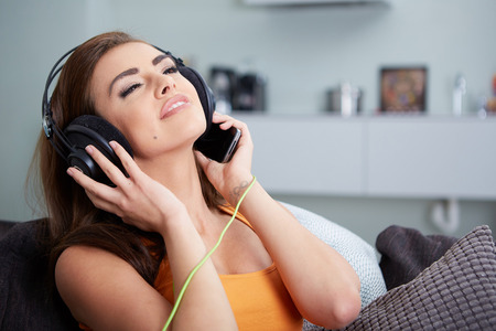 Cute smiling woman lying on couch while listening to music in bright living roomの写真素材