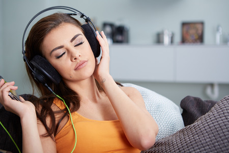 Cute smiling woman lying on couch while listening to music in bright living roomの写真素材