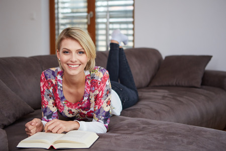 Pretty young woman enjoying reading a book at home lying on the sofa smiling in pleasure in casual clothingの写真素材
