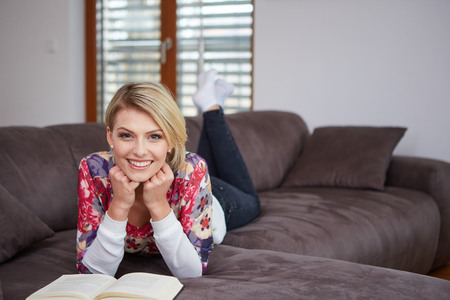 Pretty young woman enjoying reading a book at home lying on the sofa smiling in pleasure in casual clothingの写真素材