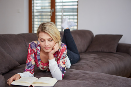 Pretty young woman enjoying reading a book at home lying on the sofa smiling in pleasure in casual clothingの写真素材