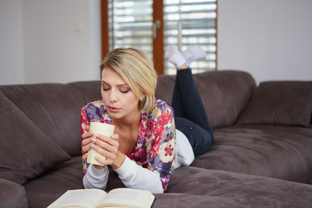 Pretty young woman enjoying reading a book at home lying on the sofa smiling in pleasure in casual clothingの写真素材