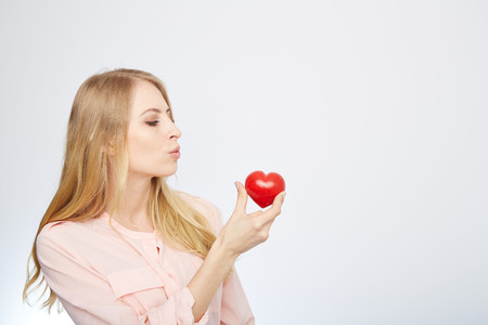 Beautiful young blond hair woman holding heart shaped valentine stone and smiling while standing isolated on whiteの写真素材