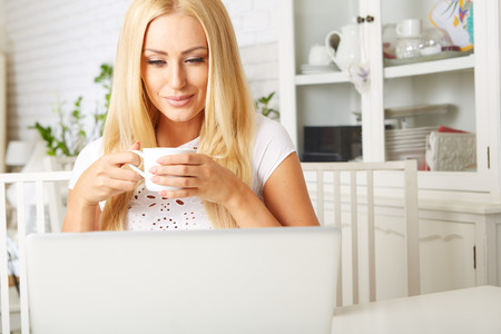 Cute woman having a cup of tea while using her laptop in her kitchenの写真素材