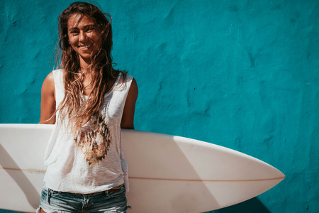 happy surfer girl with surfboard in front of blue wallの写真素材