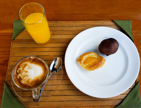 Minimal Asian breakfast setting with a glass of orange juice, cappuccino coffee and a plate with a tart and an unopened snake fruit, on a bamboo tray.の写真素材