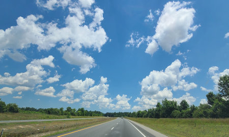 A scenic view of a highway under a bright blue sky filled with fluffy white clouds. The road is flanked by green grass and trees, creating a peaceful and inviting atmosphere.の写真素材