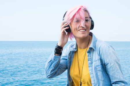 Portrait of beautiful girl sitting on rock near the sea in the headphones listening to music, with fuchsia hairの写真素材