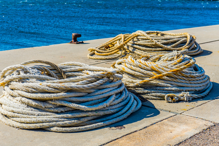 Long ropes lying on the ground at a harbor with the ocean and an anchor point visible in the background.の写真素材