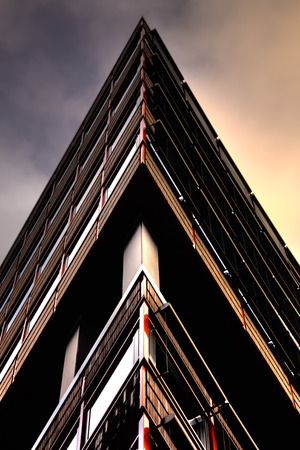 Abstract building pictured in the shape of a triangle. The sky goes from a dark shade on the left to a warmer orange color to the right side of the building. The walls have red shiny metal railings in their sides.の写真素材