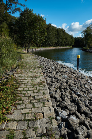 Cobblestone path near a waterway leading from a lake to a marina with a forest to the left. Vertical shotの写真素材