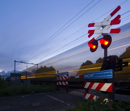 A train passing a rail crossing, surrounded by fences and barb wire on a summer evening.