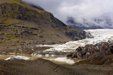 The end of the Svinafellsjokul, Iceland, with a muddy pool with icebergs and moraines, lit by a glimmer of sunlight on an overcast dayの写真素材