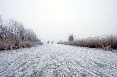 The archetypal Dutch winter on a foggy morning on a frozen canal surrounded by reed, willows, windmills and a handful of ice skaters approaching in the distanceの写真素材