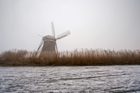 The archetypal Dutch winter on a foggy morning on a frozen canal surrounded by reed, and a windmillの写真素材