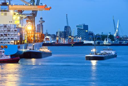 Huge container ships mooring off, with several supply trawlers around it in a commercial harbor, looking out over the Rotterdam skyline with the erasmus bridge, euromast and several high-rise office buildingsの写真素材