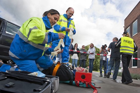 Paramedics tending to the first aid of an injured woman on a stretcher at the scene of a car crash, whilst a police woman is escorting a bystander towards the cordon tape, being filmed by a camera manの写真素材