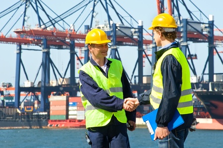 Two dockers shake hands in front of an industrial harbor with cranes and a container ship being unloaded Two dockers shake hands in front of an industrial harbor with cranes and a container ship being unloaded