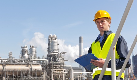 Young supervisor with a note board and pen in his hands, wearing a hard hat and safety vest in front of a petrochemical plant and refineryの写真素材