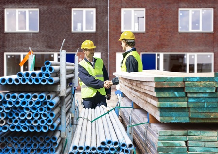 Two building contractors shaking hands behind stacks of scaffolding material, in front of a newly...