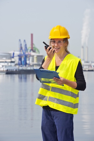 Happy female engineer communicating on a wireless radio, holding a clipboard with notes, wearing a hard hat, safety goggles and a reflective vest for safetyの写真素材