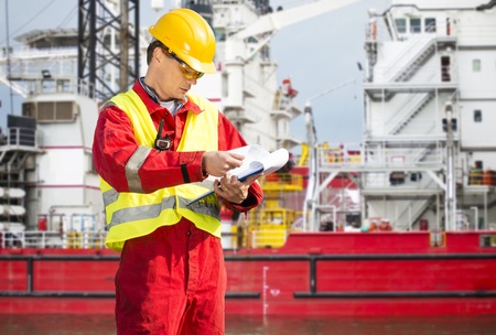 Safety officer, standing in front of a huge industrial platform, wearing overalls, a hard hat,...
