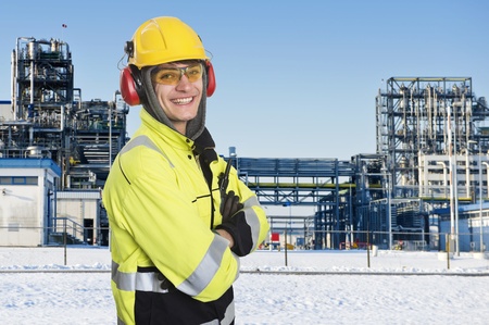 Industrial worker, posing in front of the main gate of a large chemical plant. Wintertime. Happy engineer, wearing all safety clothing necessary, such as hearing protection, a hard hat, chemical resistant reflective coat, gloves and safety glasses. Proudlの写真素材