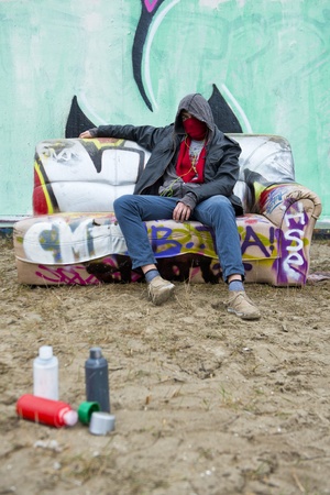 Hooded figure of a toughly posing grafitti artist, slouching on a couch in front of a grafitti clad concrete wall, with a few spray paint cans, out of focus, in the foregroundの写真素材