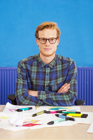 Cartoonist, sitting with his arms crossed behind his desk in front of several sketches, drawings and illustrations, littered with felt tip markersの写真素材