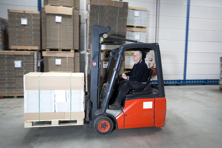 Forklift driving through a nice and clean warehouse. The experienced driver is transporting a pallet full of cardboard boxes.の写真素材