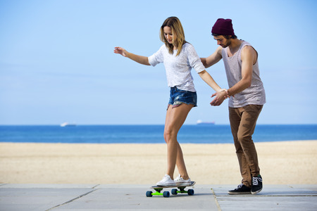 Young man gives his girlfriend her first skateboarding lesson on a seaside boulevard, during a lovely, calm summer afternoon.の写真素材