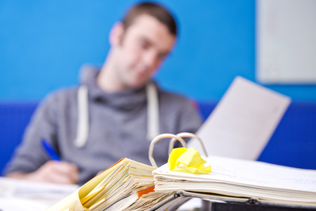 Realistic image of Paperwork, with a folder, filled with documents on a desk, with a young man behind it, reading from a heet of paper, and taking notesの写真素材