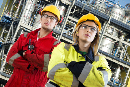 Two security engineers at their jobs, posing confidently in front of a large chemical installation with tanks, valves, tubes and safety structures Two security engineers at their jobs, posing confidently in front of a large chemical installation with tanks, valves, tubes and safety structures