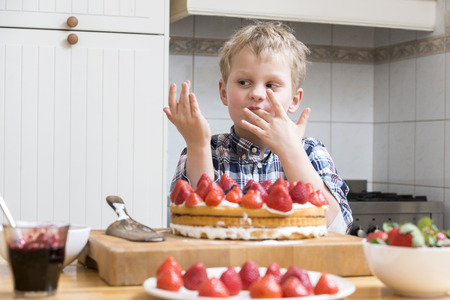 Cute boy licking his fingers with batter, behind a kitchen counter, in behind several strawberry layered cakes and piesの写真素材