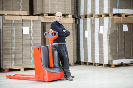 Full length portrait of confident worker standing arms crossed while leaning on handtruck at...