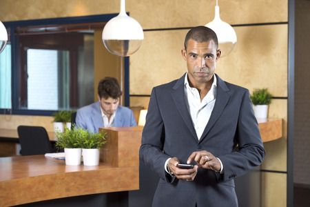 Portrait of confident businesswoman holding cellphone while receptionist working at desk in officeの写真素材