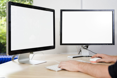 Two computer monitors with a white screen on a desk, with a man's hands on a keyboard in an office,...