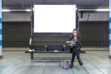 A blank billboard at an underground railway station with a woman sitting on a bench underneath, reading a bookの写真素材