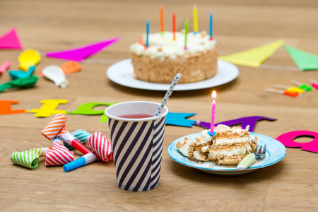 Happy birthday party still life with various garlands and objects, including a plate with cake and a candle and a paper cup with lemonade on a wooden surfaceの写真素材