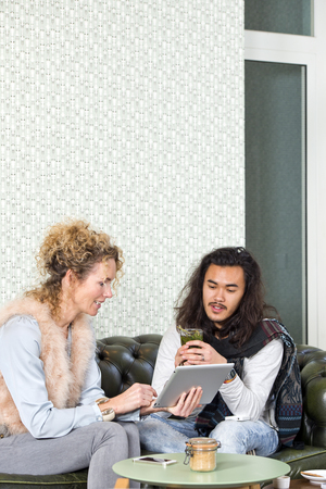 Two friends, sitting on a leather couch in the coffee corner of a lounge bar sharing photos or other information, displayed on an electronic tablet with eachotherの写真素材