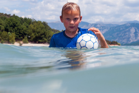 Young boy with a swimming shirt, protecting him against UV radiation, playing with a small ball in a lakeの写真素材