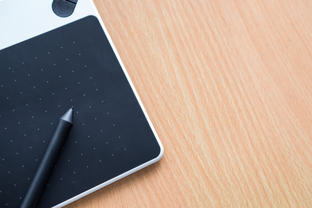 The stylus pen and pad are placed on the wooden desk. The stylus is black matching with the black...