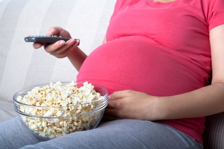 Pregnant woman sit and eat popcorn on sofa, watching televisionの写真素材