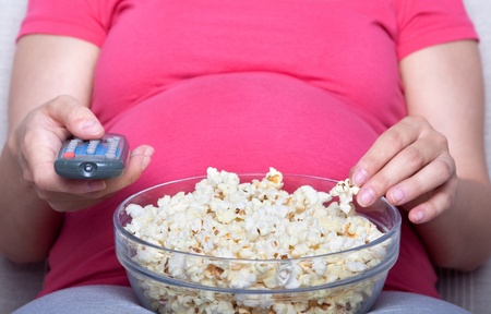 Pregnant woman sit and eat popcorn on sofa, watching televisionの写真素材