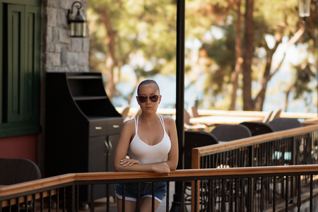 Attractive young lady in white swimsuit touching hair while standing near wooden column on resortの写真素材