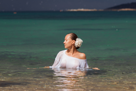 Sensual young female in wet blouse standing in clean sea water and looking awayの写真素材