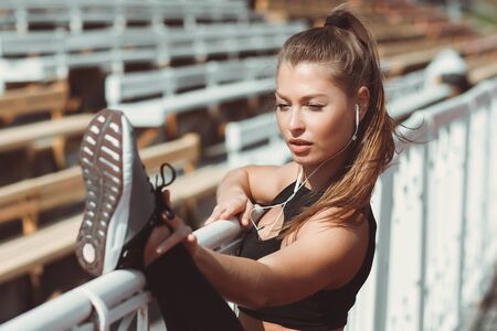 Side view of fit athletic female in sportswear and with headphones warming up while leaning on fence on urban sports groundの写真素材