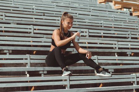Athletic woman in sportswear and earphones listening to music and drinking water from bottle while sitting on bleachers on athletic fieldの写真素材