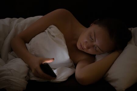 Attractive young female lying in comfortable bed late at night and using mobile phone before sleep