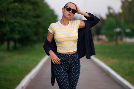 Attractive calm slim female in yellow and white striped shirt and blue with white polka dots pants in composition with unbuttoned blue jacket standing with hands in pockets and looking away against blurred summer parkの写真素材